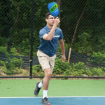 A pickleball player prepares to hit the ball with a blue and green "SLK" paddle on an outdoor court.